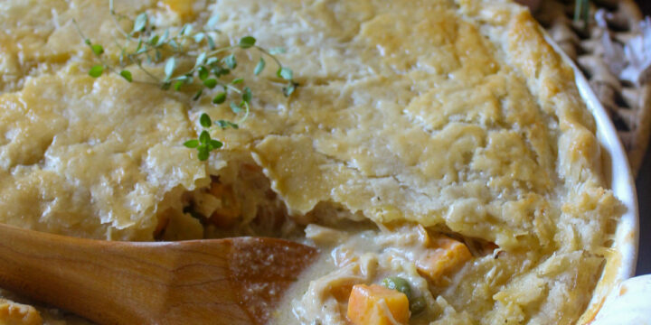 A white baking dish of chicken veggie pot pie with a flakey crust and a wooden spoon breaking into the filling.