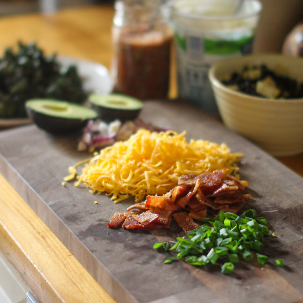 A cutting board of loaded baked potato bar toppings like shredded cheese, bacon, scallions, avocado and salsa.
