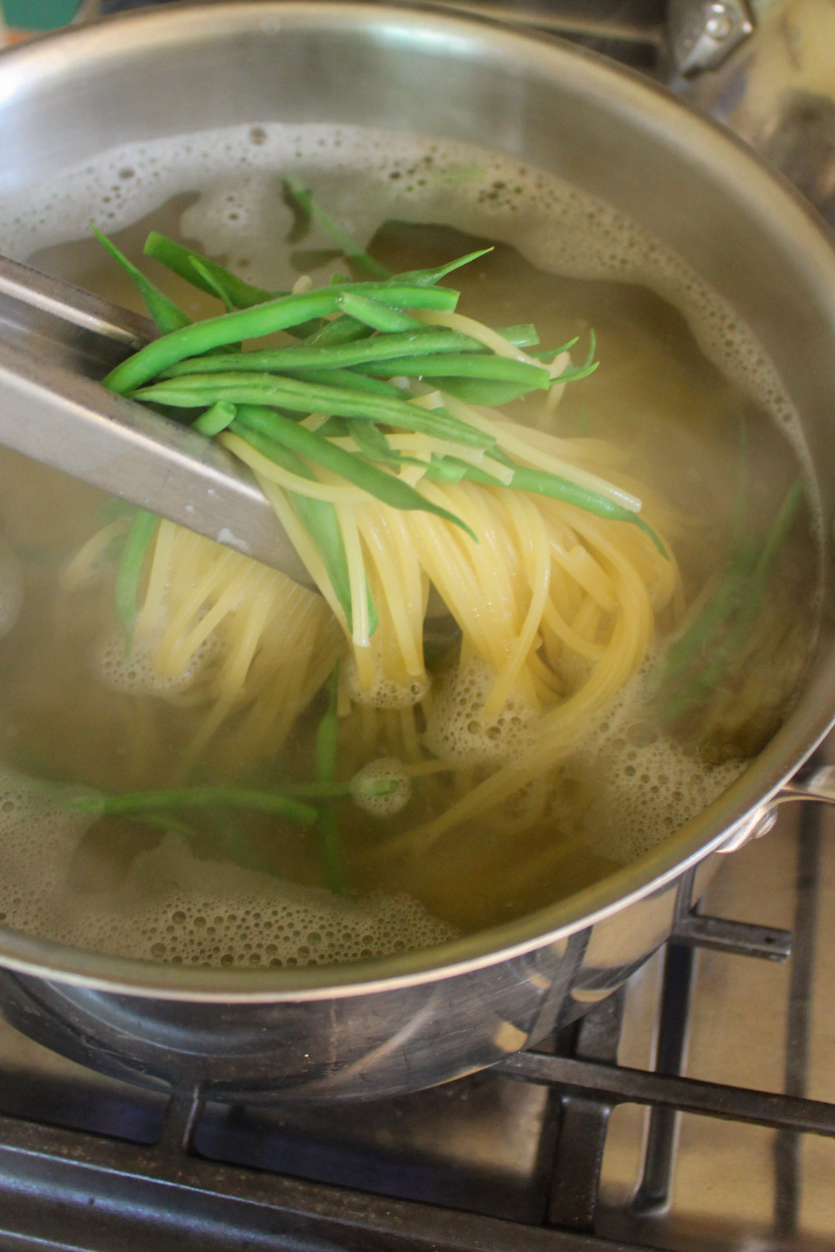 Pasta and green beans boiling together in a pot lifted with tongs.