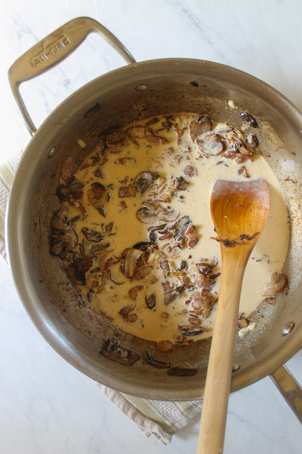 A skillet with bacon mushroom cream sauce and a wooden spoon.