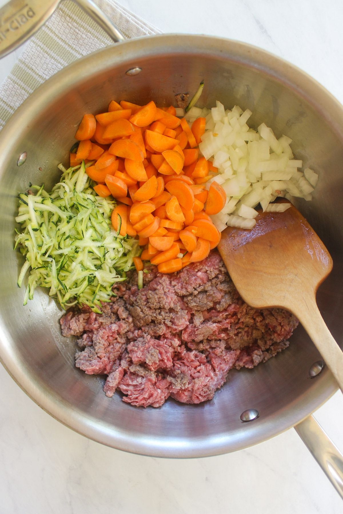 Ground beef browning on one side of a skillet with vegetables cooking on the other side.