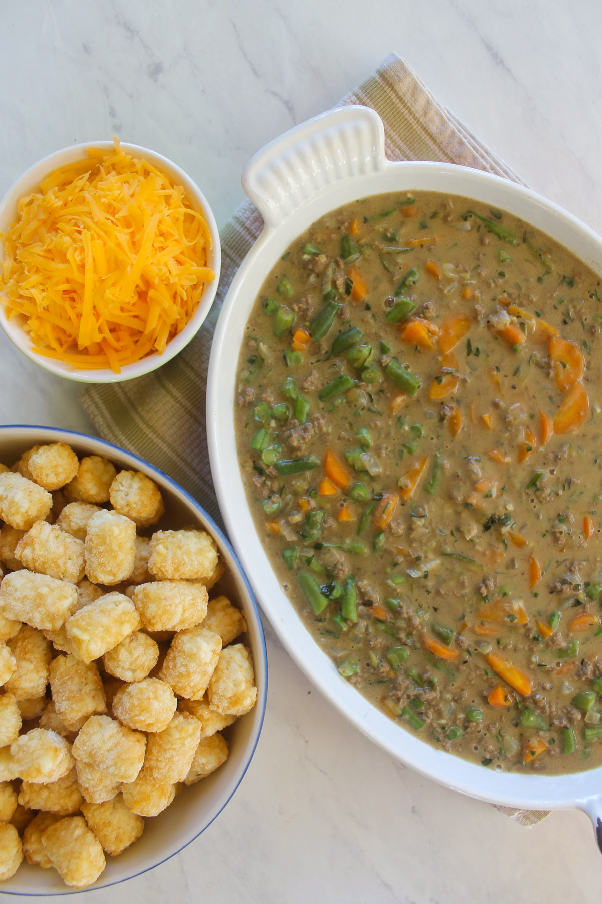 A casserole dish of tater tot casserole filling next to a bowl of cheese and frozen tater tots.