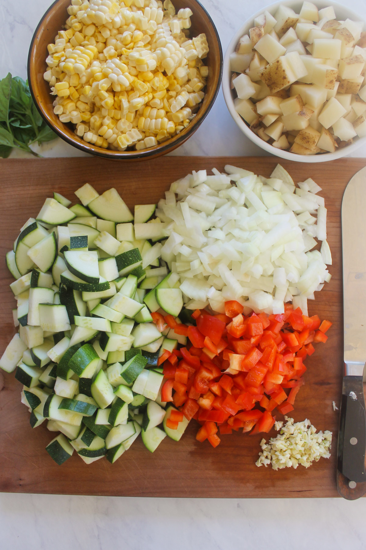 Chopped vegetables on a cutting board with bowls of potatoes and corn cut off the cob.