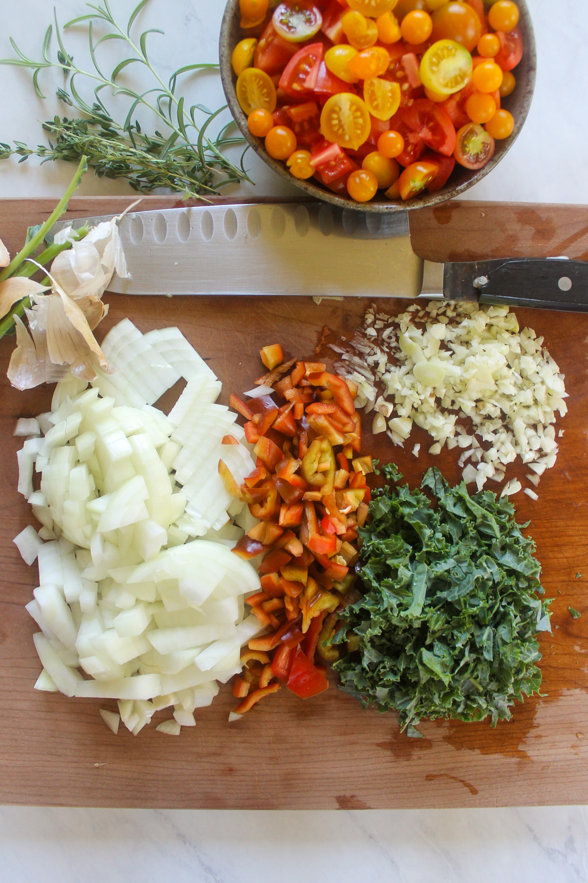 Chopped onion, red pepper, kale and garlic on a cutting board with a bowl of fresh tomatoes.