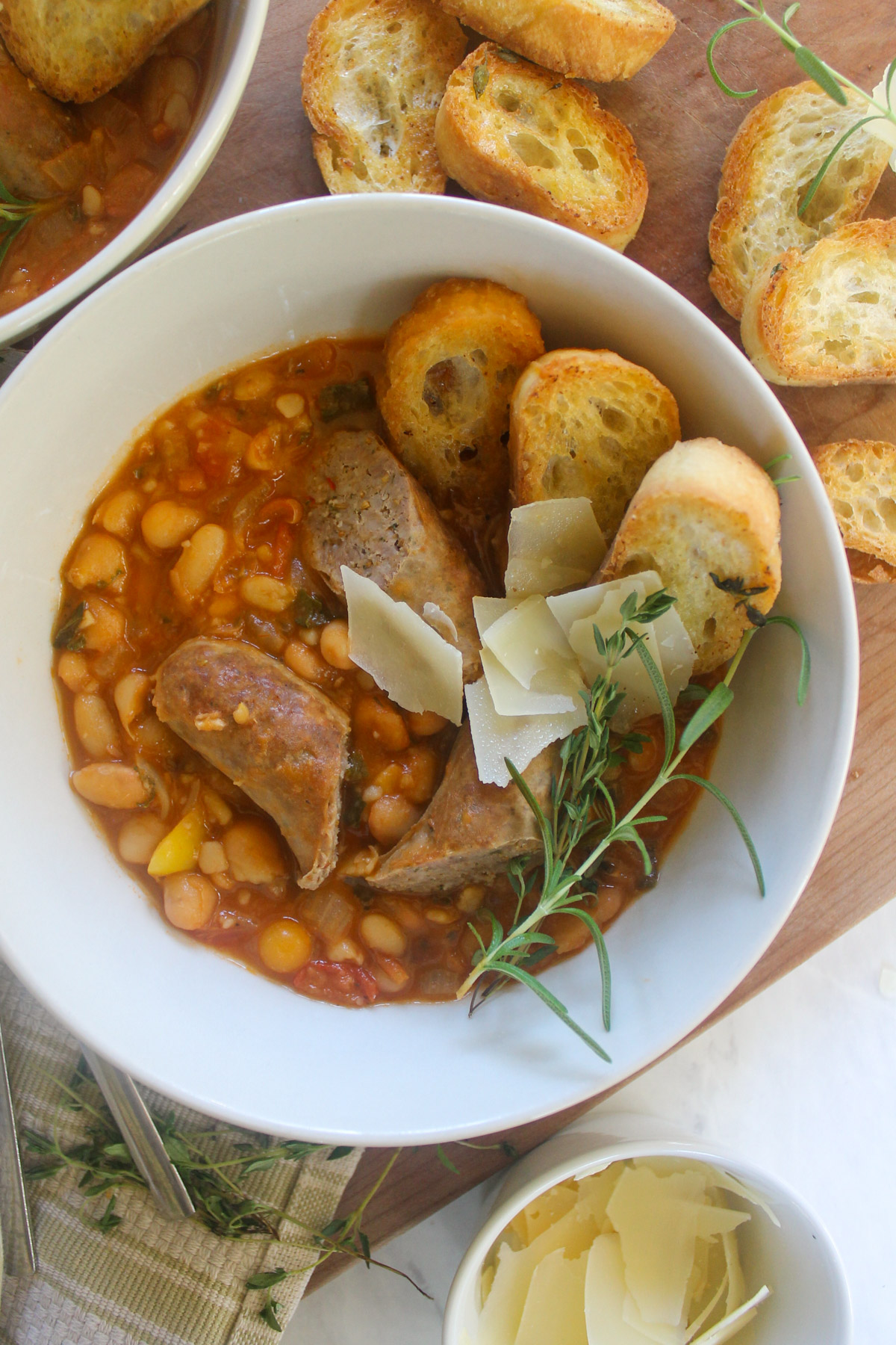 A bowl of sausage white bean cassoulet with baguette slices and Parmesan cheese.