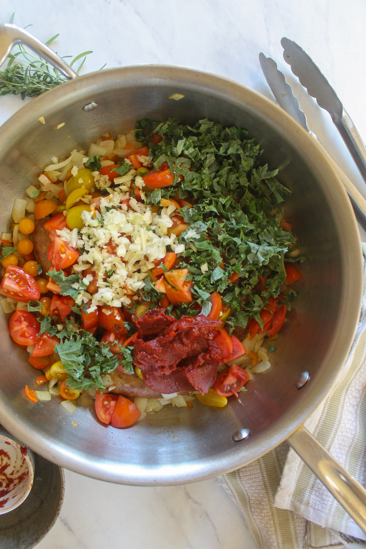 Adding fresh tomatoes, kale, garlic and tomato paste to a skillet of sausage.