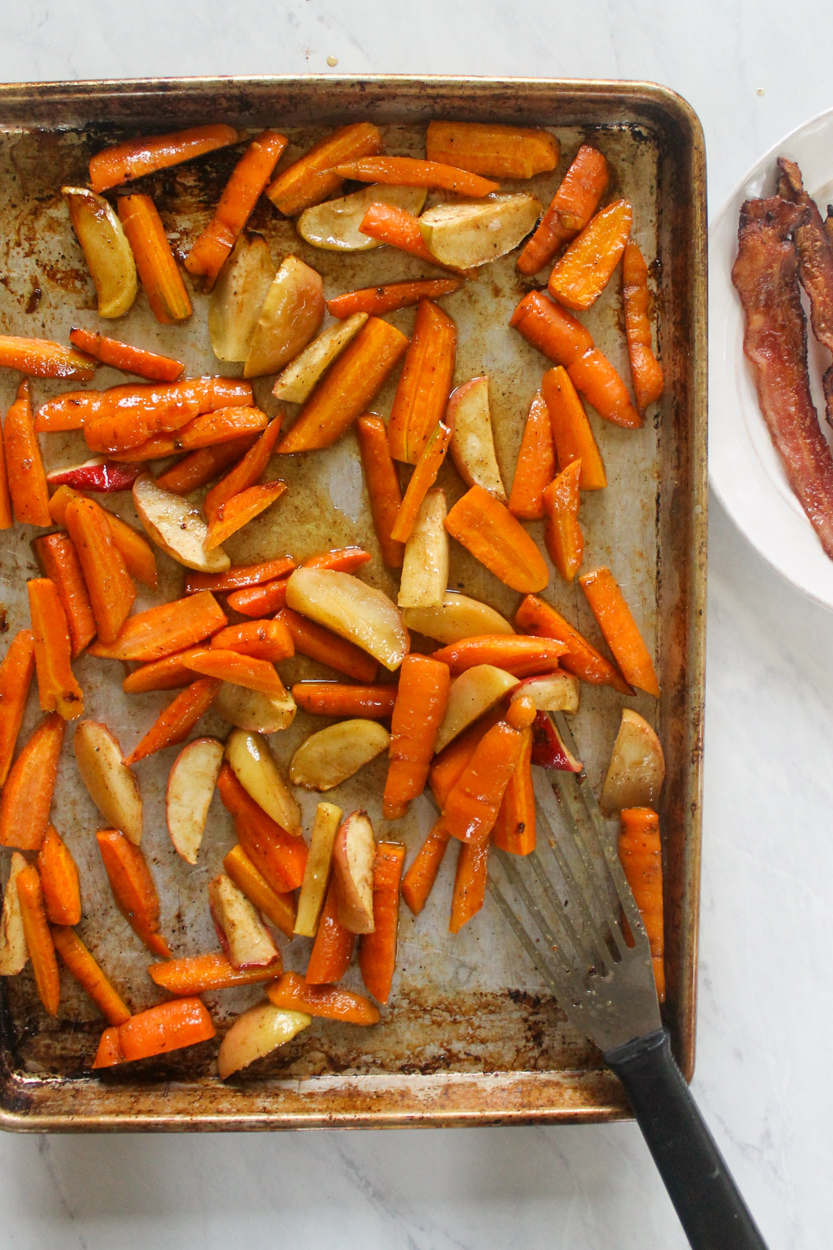 Roasting carrots and apples on a pan, bacon on a plate.