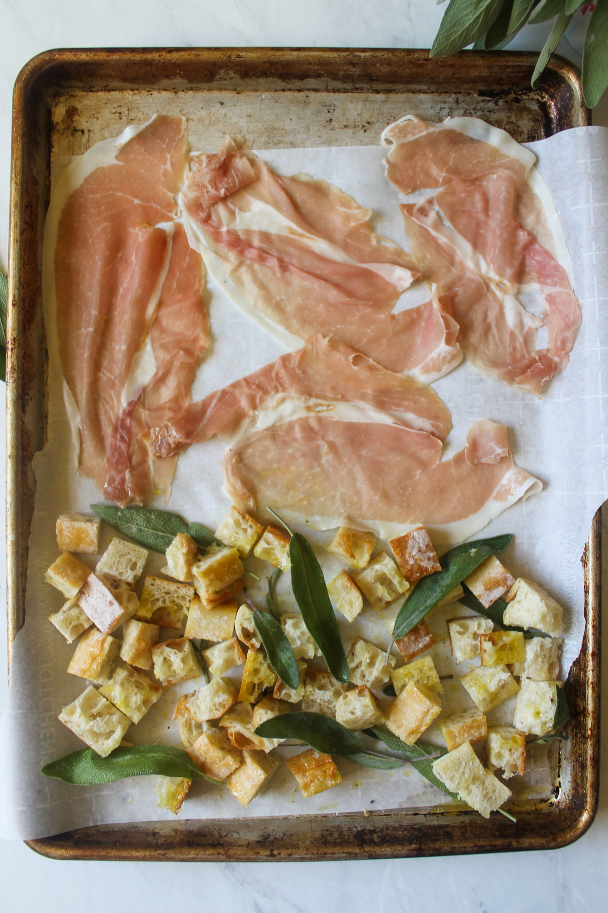 A sheet pan with slices of prosciutto and cubed bread with sage leaves.