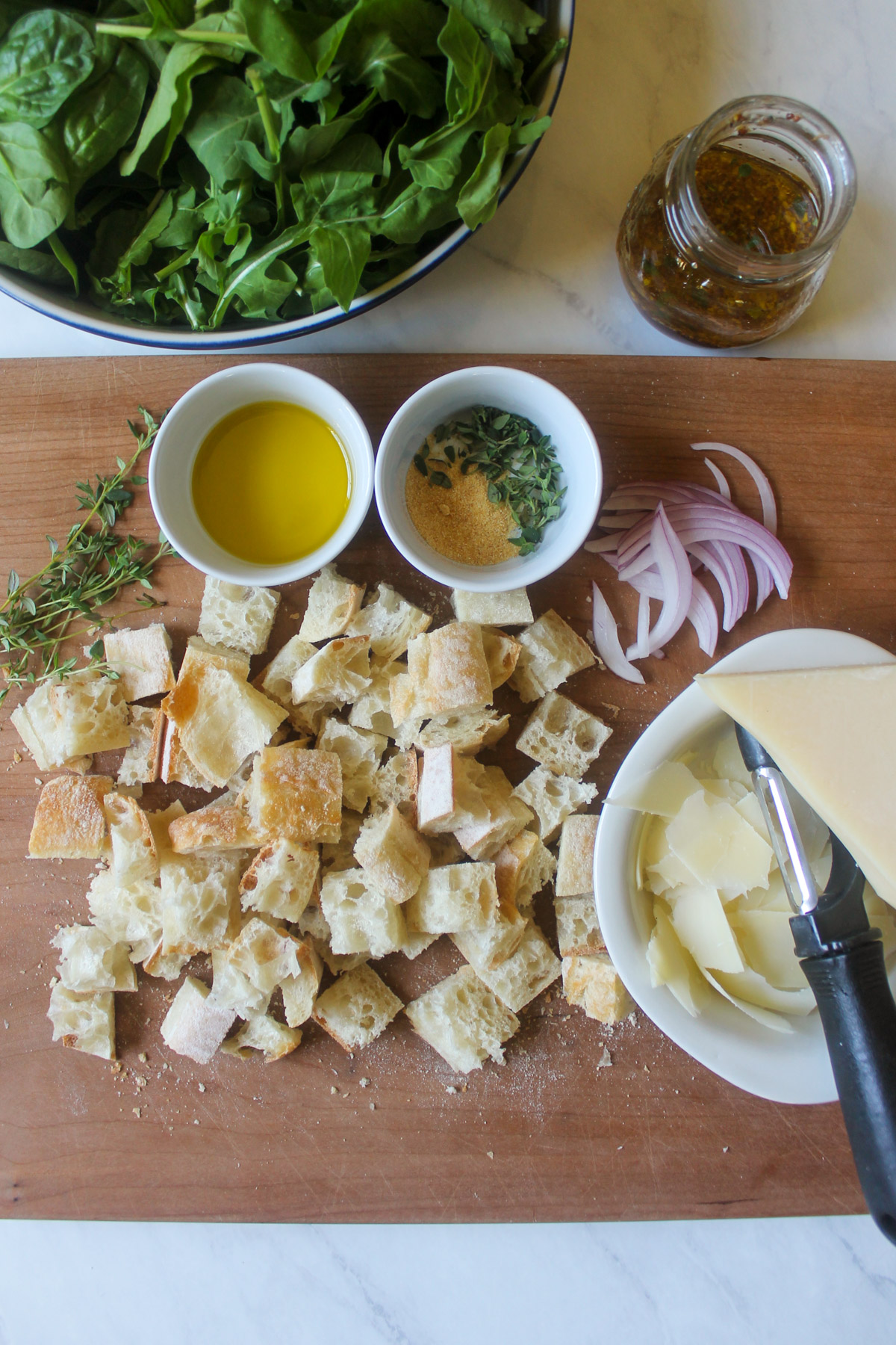 A cutting board with cubed bread, Parmesan cheese shavings, red onion slices, and a bowl of lettuce greens.