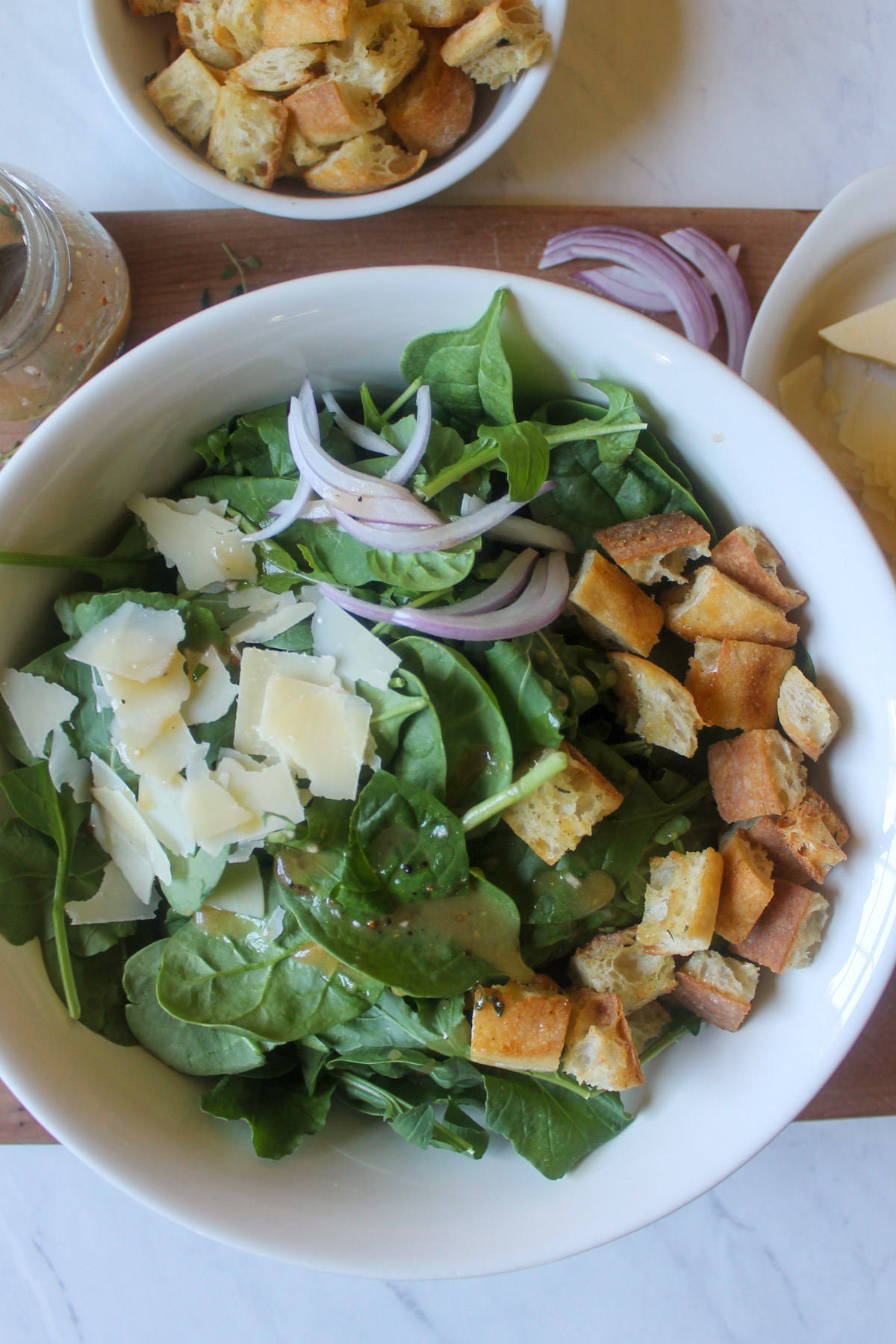 A bowl of spinach arugula salad ingredients with croutons, red onion, and Parmesan.