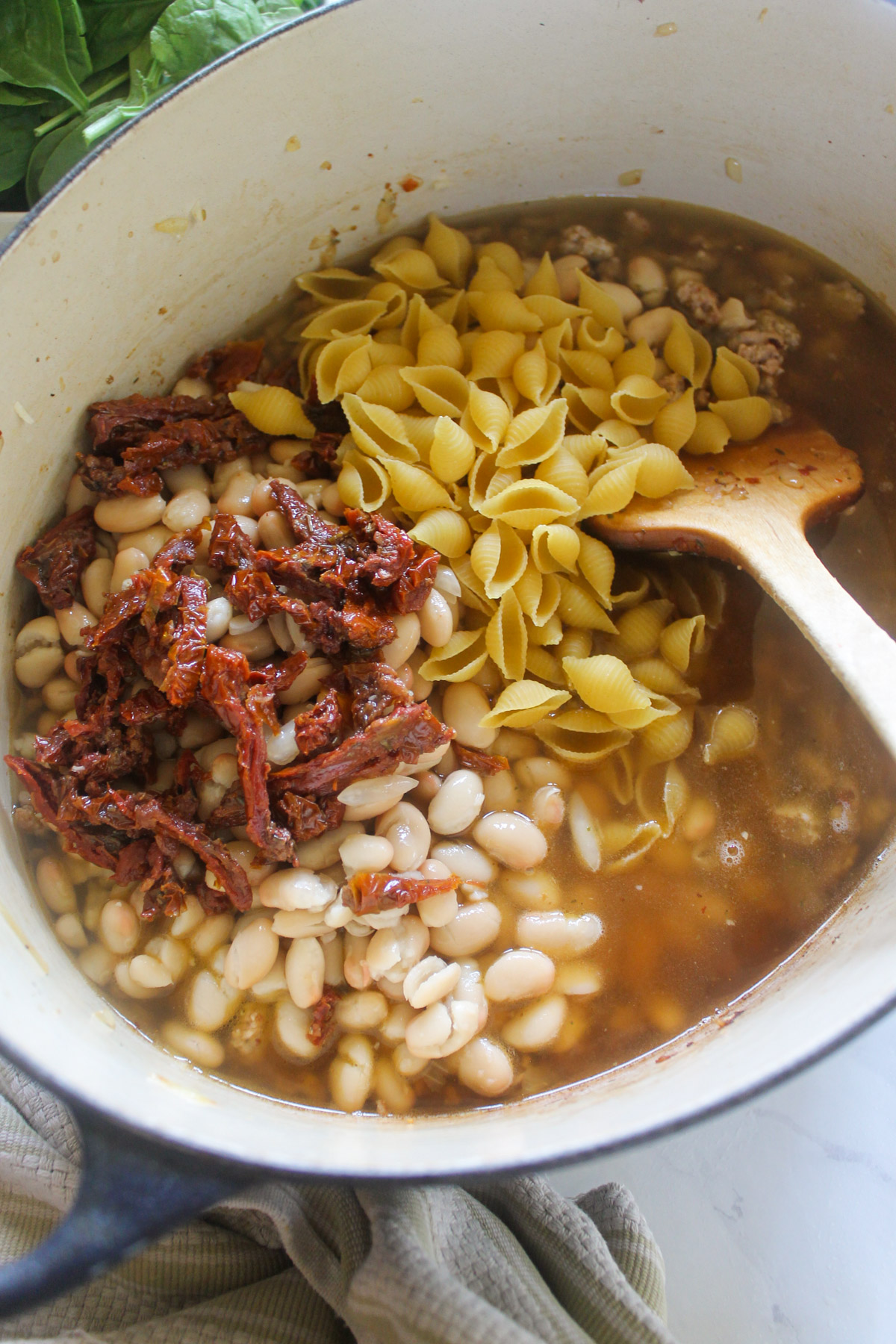 Adding dry pasta shells, white beans and sun-dried tomatoes to a pot of soup.