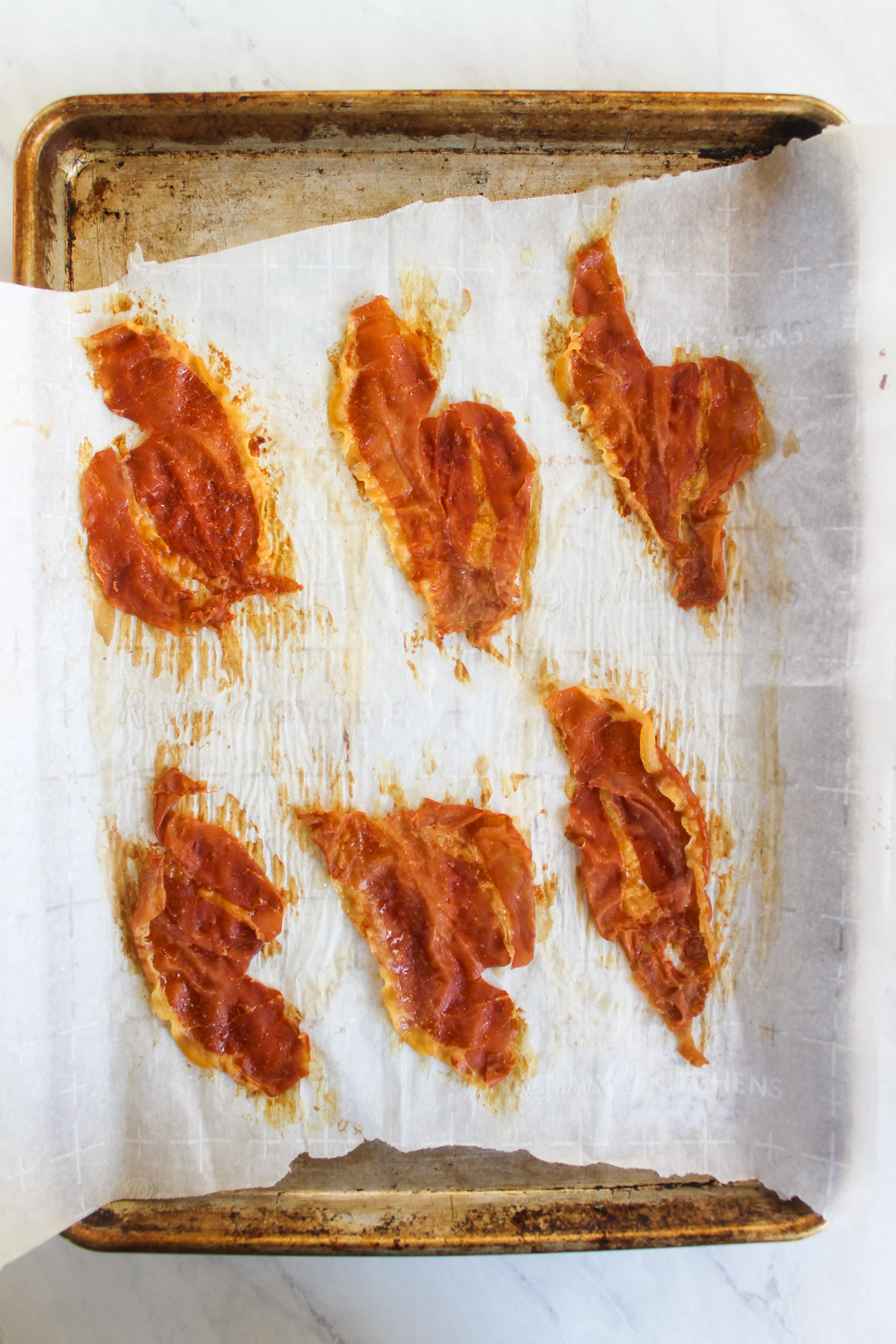 Six slices of baked crispy prosciutto on a baking sheet.