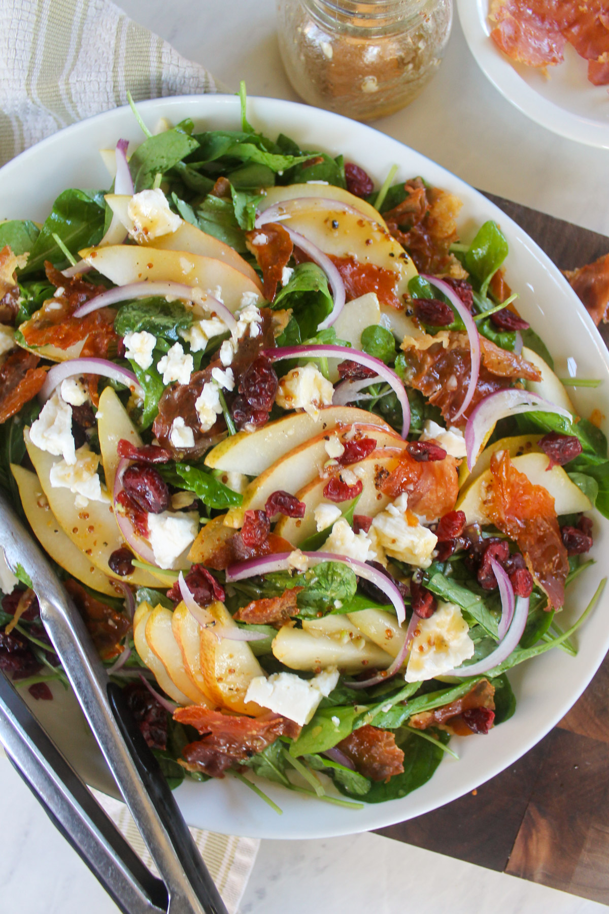 A platter of pear arugula salad with crispy prosciutto and tongs.