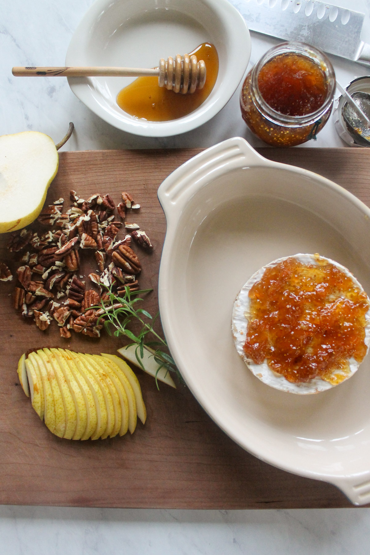 A round brie in a baking dish spread with fig jam, next to sliced pears and pecans.