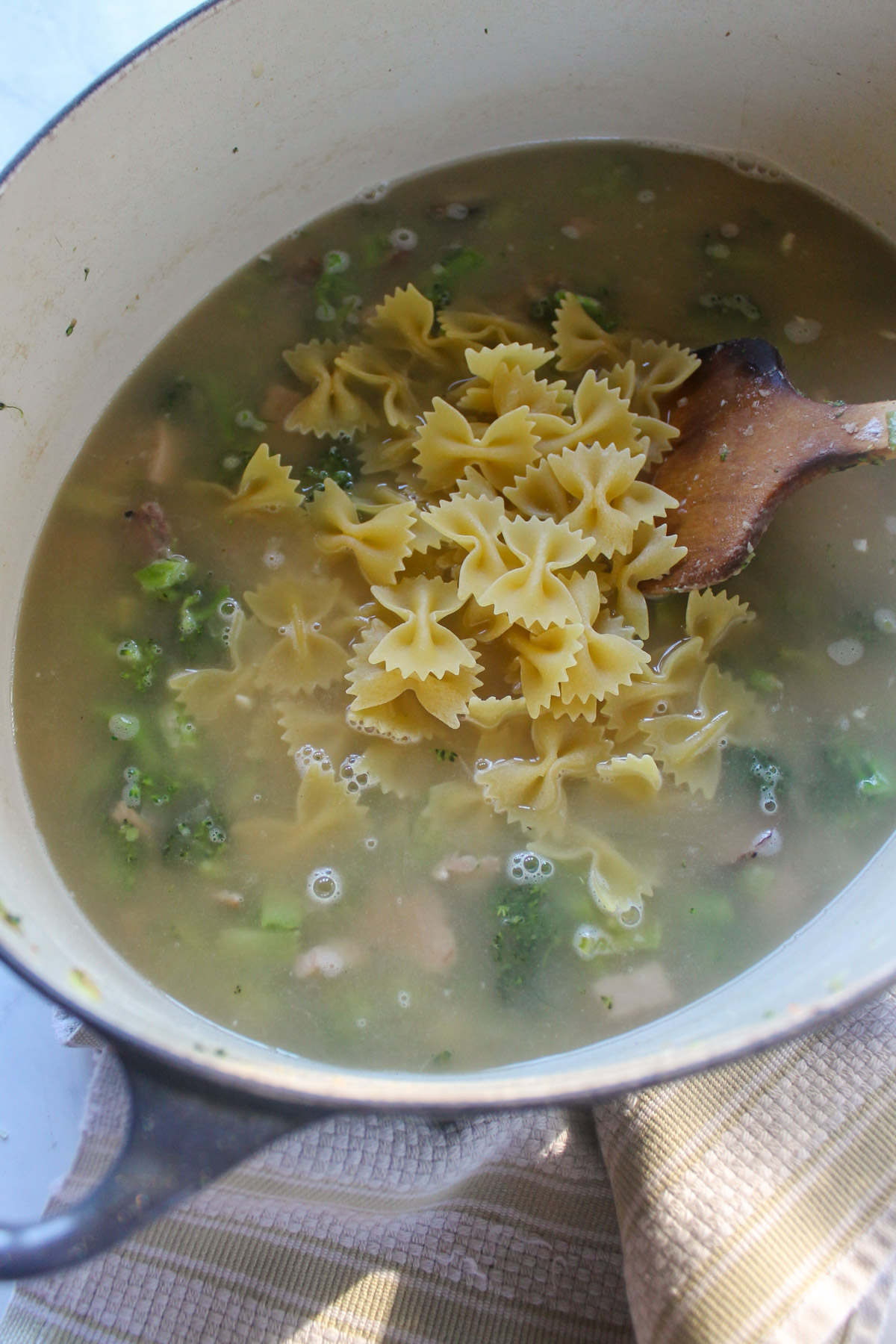 Adding chicken stock and dry bowtie pasta to a soup pot with broccoli.