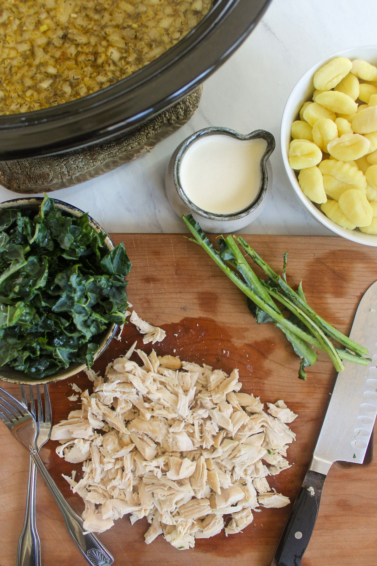 Shredded cooked chicken on a cutting board with kale, gnocchi and heavy cream in bowls.