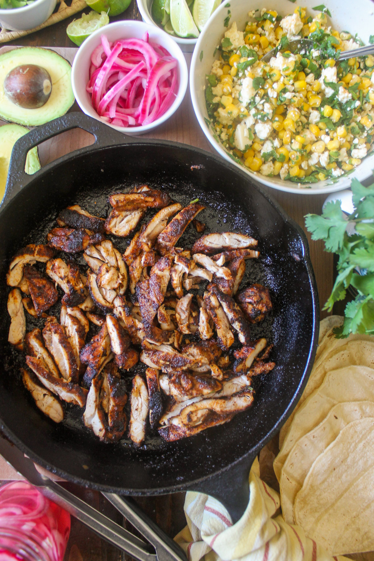 Sliced blackened chicken in a cast iron skillet with a bowl of corn salsa, pickled red onions, cilantro and avocado.