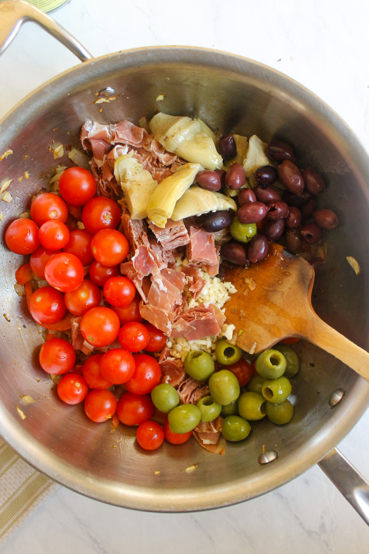 Adding cherry tomatoes, olives, prosciutto and artichokes to a skillet.