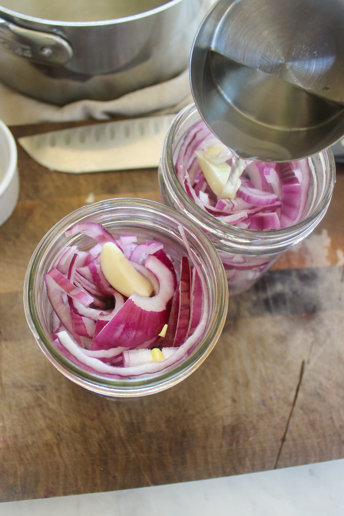 Pouring vinegar brine into two jars with red onion slices and garlic.